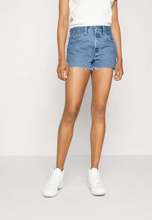 Person iført blå denimshorts med flossede kanter, hvide sneakers og en hvid top, stående mod en ensfarvet baggrund.