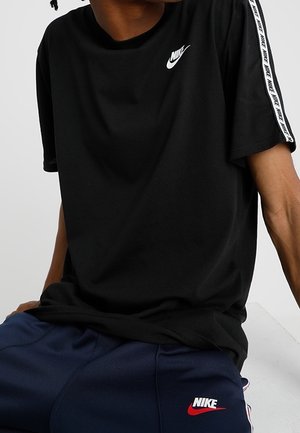 Osoba ubrana w czarną koszulkę Nike z białym logo i paskowanymi rękawami oraz granatowe spodnie Nike z czerwonym i białym logo, siedząca na neutralnym tle.