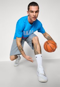 Mėlyna marškinėliai su užrašu "UNIQUE", pilkos šortai, balti sportbačiai ir krepšinis. Tatuiruotės matomos ant rankų ir kojų. Neutralus fonas.