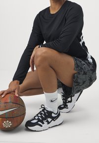 Haut noir à manches longues, short noir à motifs, chaussettes blanches Nike et chaussures noires Nike. Ballon de basket en main. Design angulaire et athlétique.