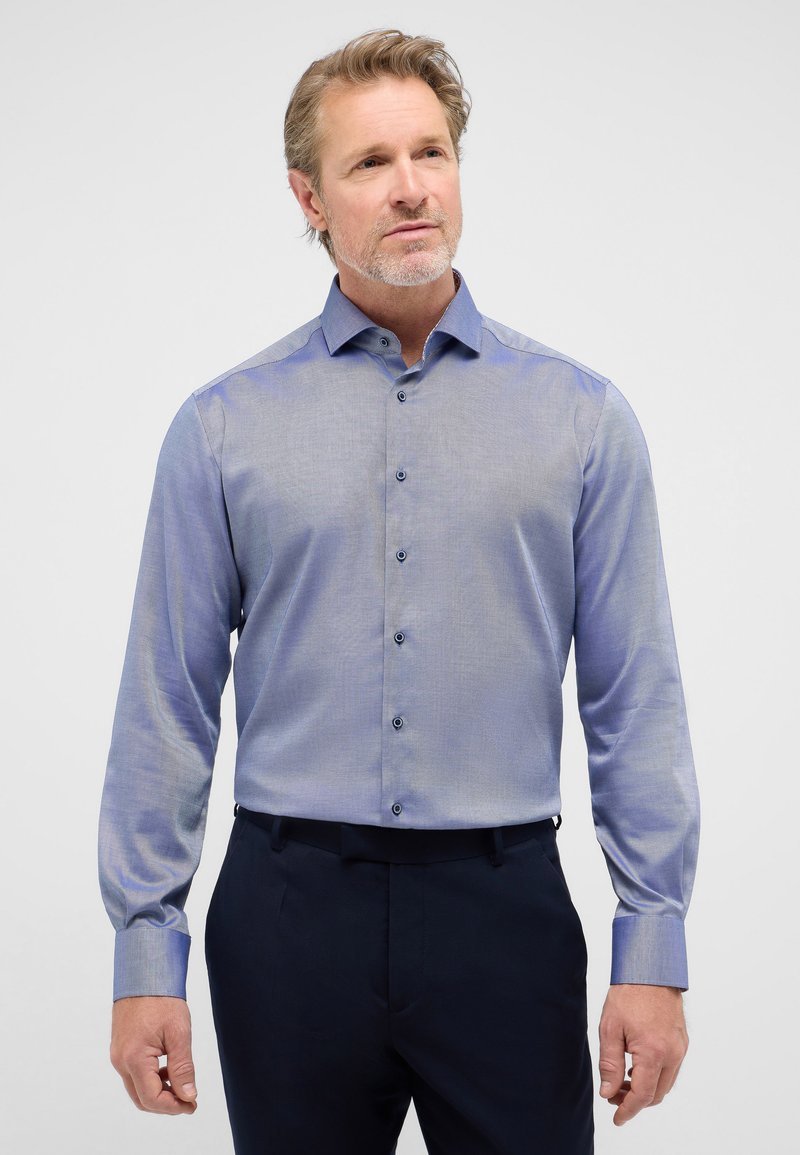 Lichtblauw overhemd met lange mouwen en een subtiel ruitpatroon, met button-down kraag en blauwe knopen, gecombineerd met donkere broeken.