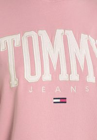Ljust rosa sweatshirt med stora vita broderade texter "TOMMY" och "JEANS", med en liten grafik av en marinblå, vit och röd flagga längst ner.