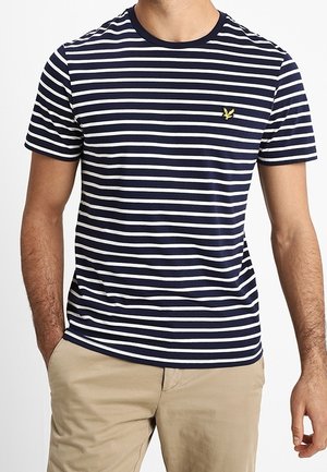 Homme portant un t-shirt à manches courtes rayé horizontal bleu marine et blanc avec un petit logo oiseau jaune, associé à un pantalon beige, mains dans les poches.