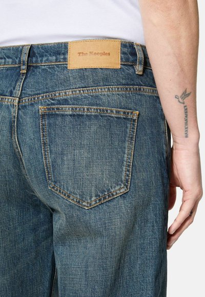 Person wearing blue denim jeans with back pocket and leather brand label "The Kooples" above the pocket, arm tattoo visible.