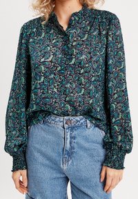 Chemise à manches longues en tissu foncé ornée de motifs floraux verts délicats, avec un col mandarin et des manches smockées, associée à un jean en denim clair.