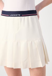 Plisserad tenniskjol i krämfärgad tyg, med midja i marinblått med "LACOSTE"-logotyp och röda och vita detaljer.