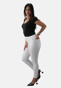 Weiße enge Jeans mit hoher Taille, kombiniert mit einem figurbetonten schwarzen T-Shirt. Schwarze Absätze mit dekorativen Akzenten und schmalen Knöchelriemen.