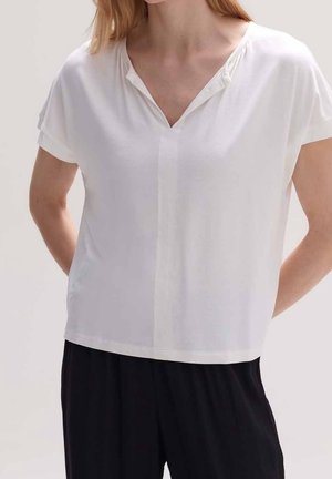 Femme portant un chemisier blanc à manches courtes et col en V, ample, et un pantalon noir, debout les mains derrière le dos sur un fond uni.