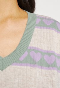 Close-up van een sweaterhals met geribbelde mintgroene rand en een patroon van paarse harten op een beige en mint achtergrond.