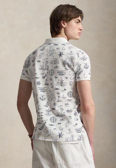 Νέος άνδρας που φοράει λευκό πόλο με μαύρα ναυτικά σχέδια, όπως ιστιοπλοϊκά, άγκυρες, πυξίδες και ναυτικά σύμβολα, κοιτάζοντας πλάγια.