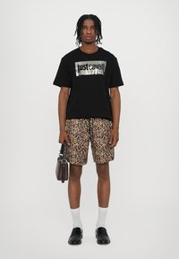 Zwarte T-shirt met metallic "justcavalli" logo, gecombineerd met luipaardprint shorts. Witte sokken en zwarte schoenen, met een bruine tas.