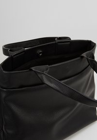 Sac cabas en cuir noir texturé avec ouverture supérieure et deux bretelles, montrant la doublure intérieure et une fermeture à pression magnétique.