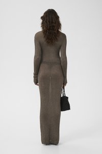 Žena s dlouhými vlnitými vlasy, oblečená v přiléhavých hnědých pletených šatech s dlouhým rukávem, která drží černou korálkovou kabelku, stojí před bílým pozadím.