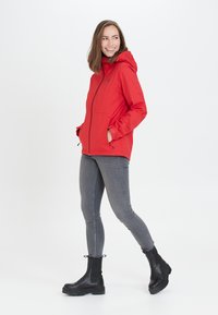 Veste rouge imperméable avec capuche, fermeture éclair à l'avant, et deux poches latérales, associée à un jean skinny gris et des bottines noires. Fond uni.