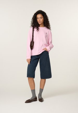 Woman with curly hair wearing a pink long-sleeve shirt, knee-length dark denim shorts, gray socks, brown flats, and a brown shoulder bag.