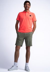 Krátký rukáv, jasně oranžová polo tričko s límečkem, kombinované s olivově zelenými kraťasy. Bíl soky a světlé tenisky dokončují outfit.