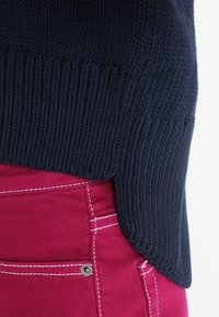 Der Saum des marineblauen Strickpullovers ist über den roten Hosen sichtbar. Der Pullover weist eine strukturierte Rippung auf; die Hose hat kontrastierende weiße Nähte und metallische Beschläge.