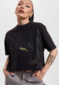 Černé zkrácené tričko z bavlny s odvážným grafickým designem, na kterém je nápis "ROCANWEAR" s různou průhledností, doplněný žlutým písmem.