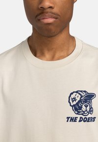Beiger Sweatshirt mit einem navyfarbenen Aufdruck eines Hundes in einer Mütze und dem Text "DIE MACHER" darunter. Weicher Stoff und Rundhalsausschnitt.