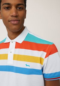 Witte poloshirt met horizontale strepen in rood, geel, turquoise en blauw, met een klein turquoise teckel-logo op de borst.