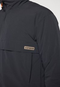 Svart softshell-jakke med glidelås foran, høy krage og oransje detaljer på glidelåsen. Har en horisontal brystlomme og logoapplikasjon.