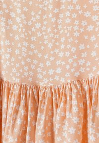 Pfirsichfarbener Stoff mit weißem Blumenmuster; hat eine geraffte Textur entlang des Saums, die eine sanfte, fließende Silhouette erzeugt.