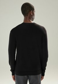 Man facing away wearing a plain black sweater and gray pants against a neutral background.