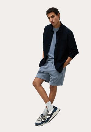 Camisa de botones azul marino sobre una camiseta gris claro, combinada con pantalones cortos gris claro y zapatillas azul marino con detalles grises y calcetines blancos.