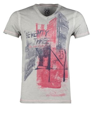 Mężczyzna w szarej koszulce z nadrukiem, czarnych dżinsach, bordowych trampkach, noszący czarną torbę na ramieniu i trzymający granatową kurtkę typu varsity.