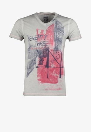 Man draagt een grijs t-shirt met print, zwarte spijkerbroek, kastanjebruine sneakers, draagt een zwarte tas op de schouder en houdt een donkerblauwe varsity-jas vast.