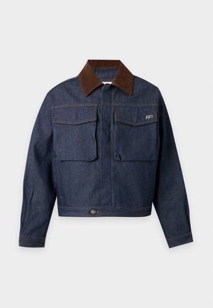 Jeune homme debout les mains dans les poches, portant une veste en jean bleu foncé avec col marron, une chemise beige, un pantalon marron à jambes larges et des chaussures bicolores.