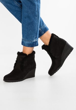 Czarne botki na koturnie z futrzanym wykończeniem, noszone z podwiniętymi niebieskimi dżinsami, na białym tle podłogi.