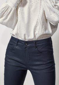 Mörkblå, åtsittande jeans med en jämn yta, femficksdesign och knäppning. Matchas med en vit blus med spetsdetaljer.