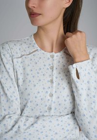 Blusa azzurro chiaro con motivo floreale, maniche lunghe, scollatura rotonda e chiusura a bottoni. Presenta rinforzi decorativi in pizzo sulle spalle.