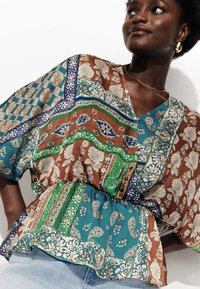 Blouse à motifs marron et teal avec un décolleté en V, une taille élastique et des manches larges. Présente des motifs floraux et de paisley sur l'ensemble.