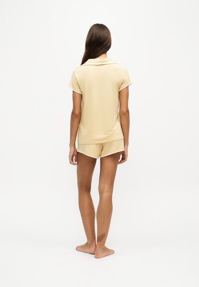 Femme aux longs cheveux bruns, pieds nus et de dos, portant un haut jaune pâle à manches courtes et un short assorti avec un bord blanc.