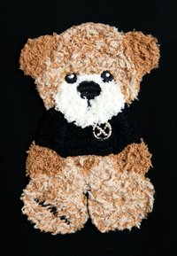 Osito de peluche con pelo texturizado en tonos marrón y crema, vestido con un suéter negro que presenta un acento dorado circular. Detalles de ojos y nariz suaves.
