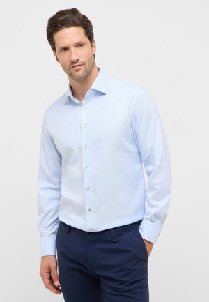 Mann mit kurzen braunen Haaren, der ein hellblaues Hemd mit Knöpfen und dunkelblaue Hose trägt, steht mit einer Hand in der Tasche.