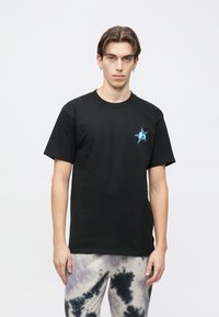 Juodas medvilninis trumpomis rankovėmis marškinėlis su mėlynu žvaigždės logotipu kairėje krūtinės pusėje. Dėvimas su pilkais ir baltais tie-dye raštu kelnėmis.