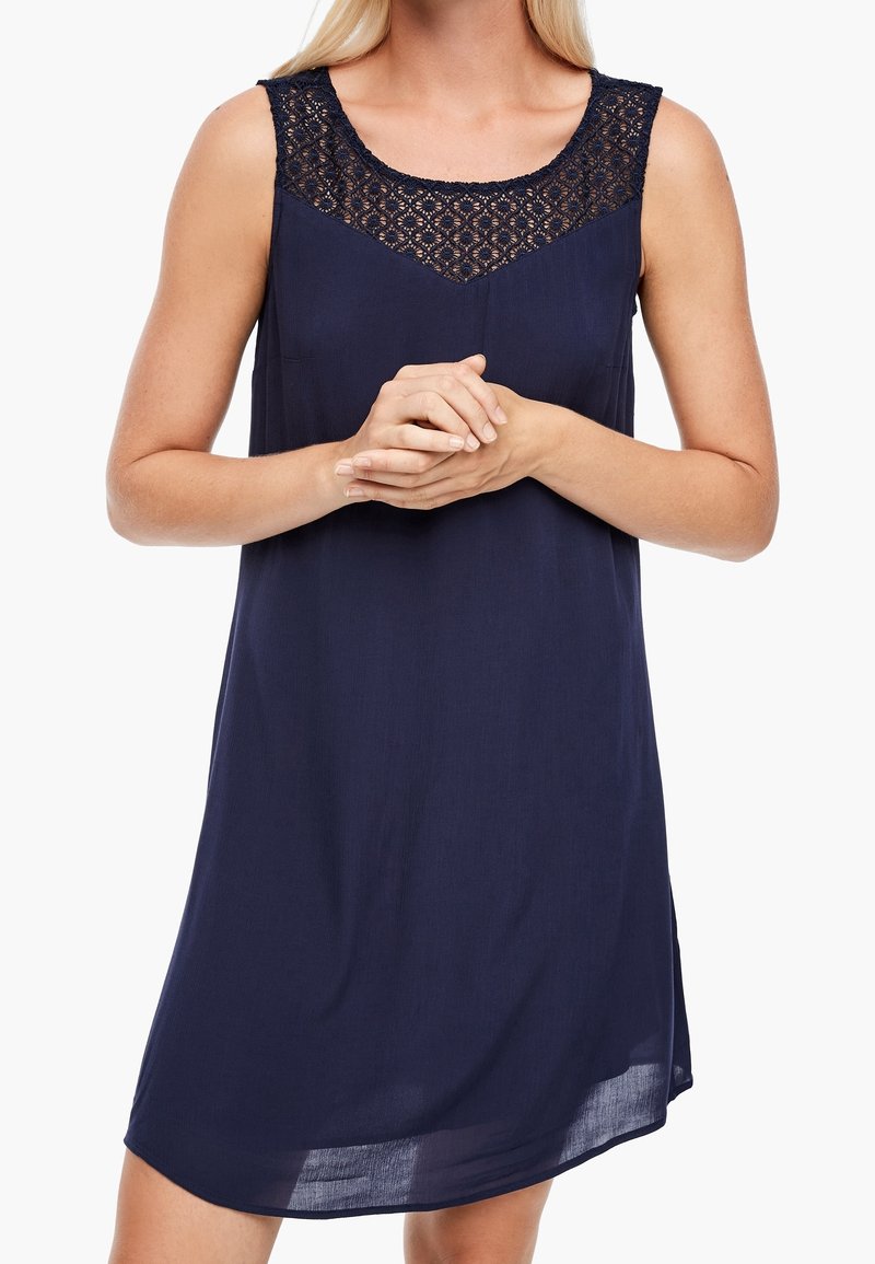 Femme portant une robe sans manches bleu marine avec un détail en dentelle au niveau de l'encolure, les mains jointes devant, sur fond blanc.