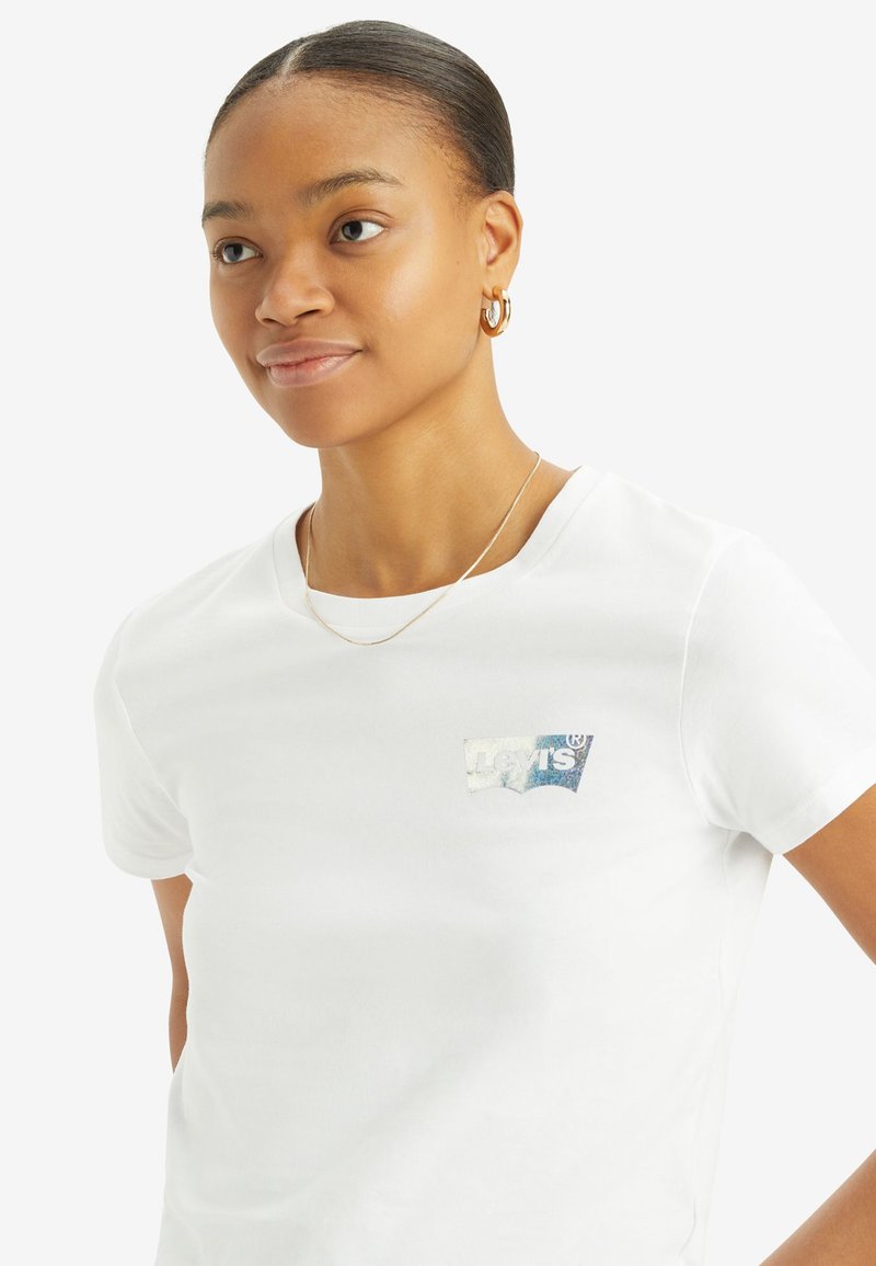 T-shirt en coton blanc à manches courtes, arborant un patch logo bleu et argent sur la poitrine gauche. Texture lisse et coupe classique.