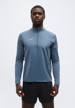 Άνδρας με μπλε αθλητική μπλούζα μακρυμάνικη με μικρό φερμουάρ στο μπροστινό μέρος και μαύρο σορτς, που στέκεται μπροστά σε απλό φόντο.