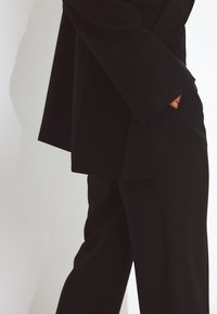 Črn usklajen kostim, ki vključuje blazer s širokimi rokavi in ravne hlače. Material se zdi gladek in lahek, vidne so tudi podrobnosti žepov.