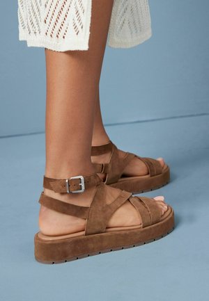 Sandali in suede marrone con una piattaforma piatta, caratterizzati da cinturini incrociati e una fibbia alla caviglia. Texture morbida e design della suola robusto.
