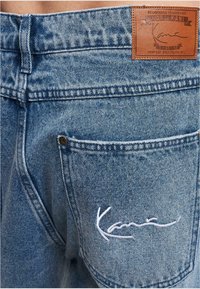 Modré džíny z denimu s texturovaným povrchem, vyznačující se koženou nášivkou značky a bílým vyšitým logem na zadní kapse.