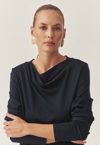 Sötét tengerészkék pajzsnyakú felső hosszú ujjal, puha textúrával és laza szabással. A modell nagy, kerek arany statement fülbevalókat visel.