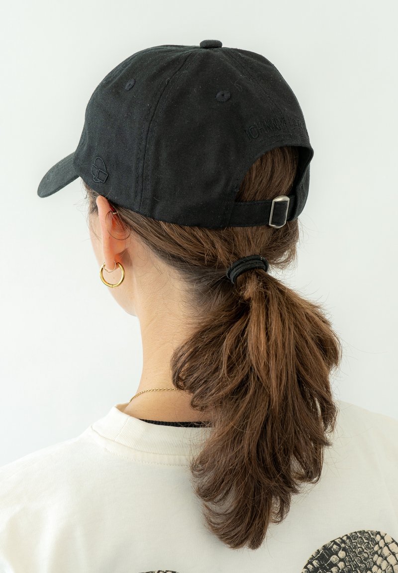 Personne aux cheveux bruns en queue de cheval portant une casquette de baseball noire et une chemise blanche, vue de dos sur un fond blanc uni.