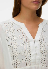 Gros plan d'une femme portant un chemisier blanc avec une broderie florale en broderie anglaise et trois boutons recouverts de tissu sur l'encolure avant.