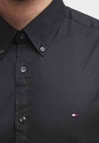 Camisa negra de botones con cuello abotonado y pequeño logo rectangular en rojo, blanco y azul en el pecho, llevada por una persona.