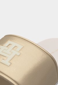 Beige Riemchensandale mit einem erhöhten weißen Doppel-T-Logo auf dem Riemen, das eine strukturierte Oberfläche und eine gebogene Kante zeigt.